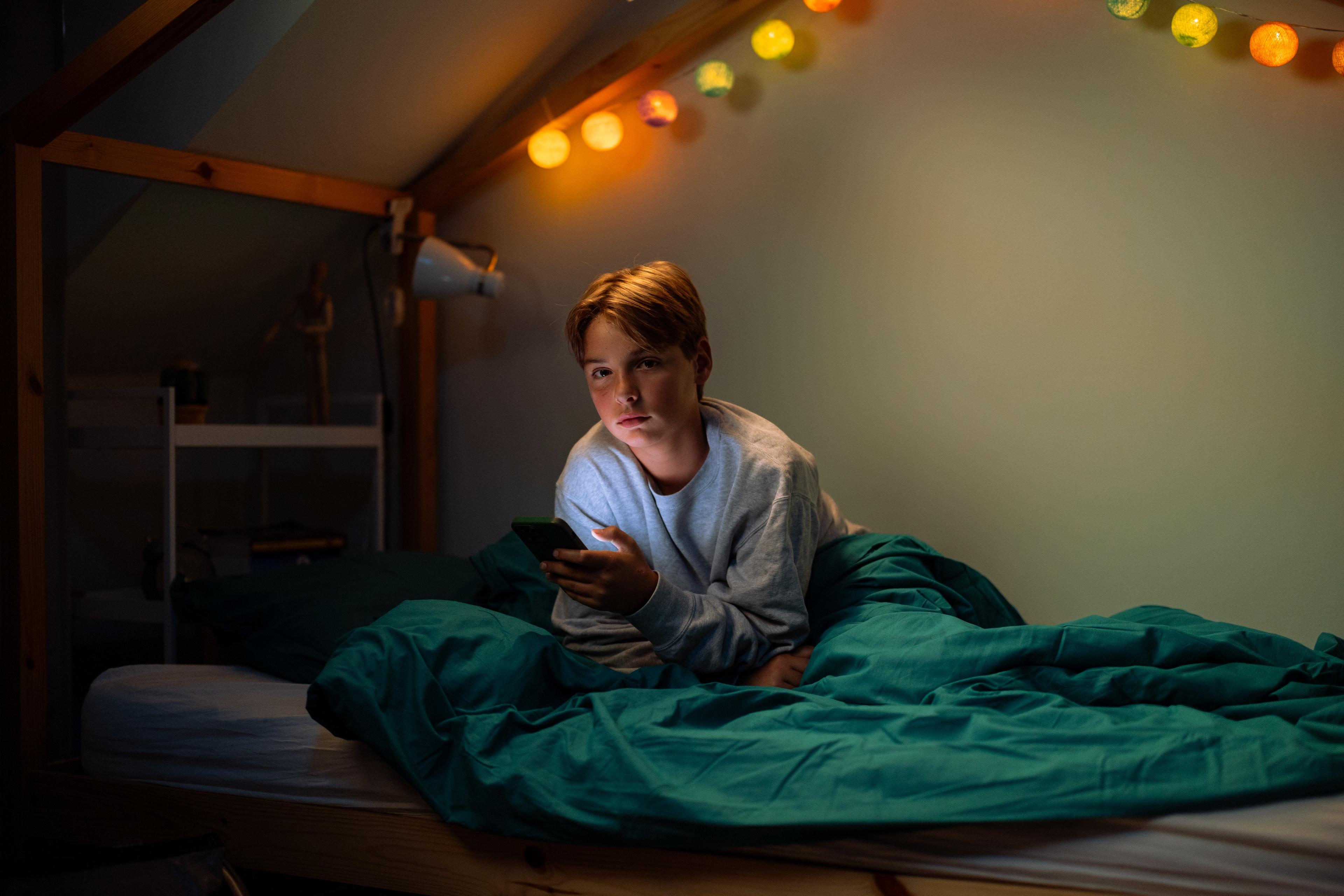 Foto van een jongen met donkerblond haar liggend op zijn bed met telefoon in hand, kijkend naar de camera.