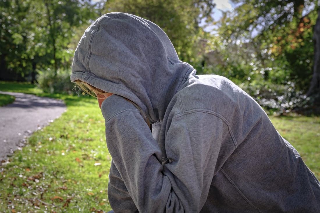 Jongen met een grijze trui op een bankje buiten, met zijn hoofd in zijn handen