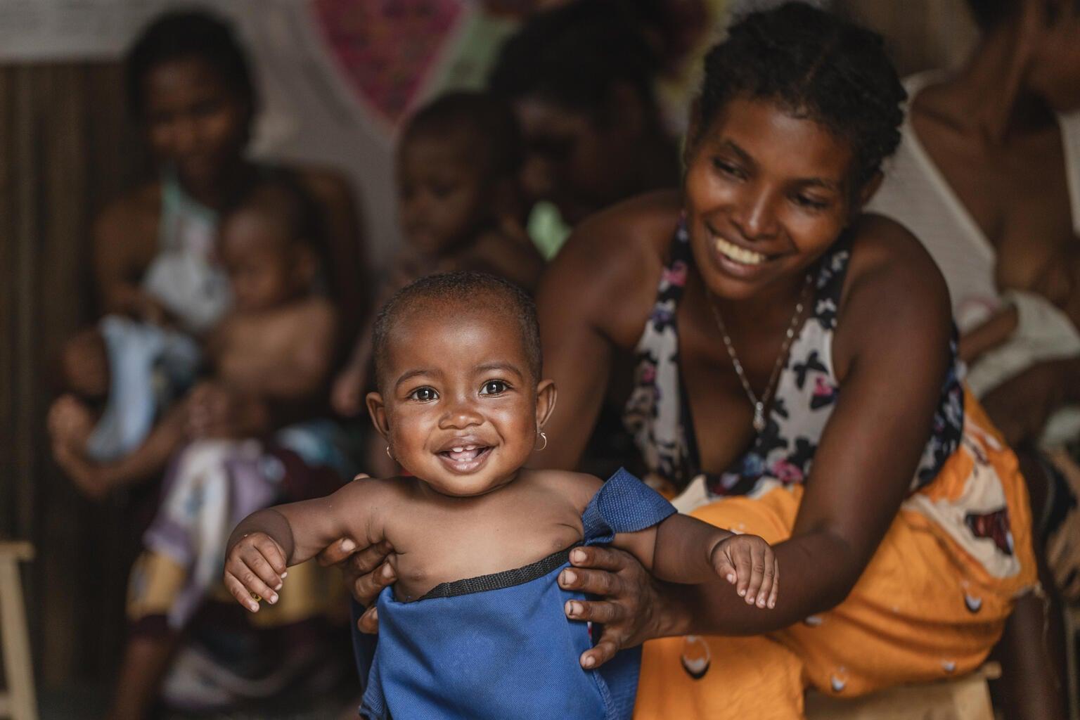 Lachend meisje uit Madagaskar wordt gewogen tijdens een medische check, moeder op achtergrond