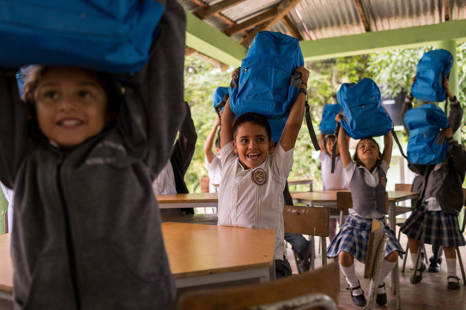 Ontheemde kinderen in een klaslokaal in Colombia houden enthousiast hun rugzakken omhoog.